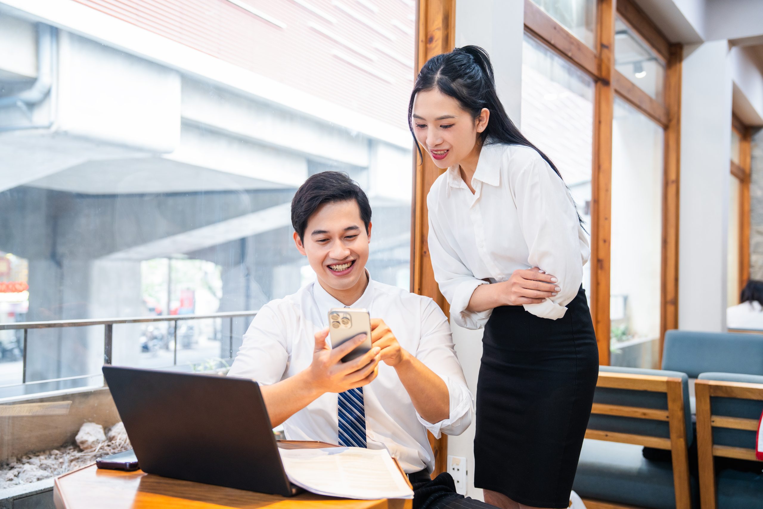 Two office colleagues look at a mobile phone in the office with laptop business discussion